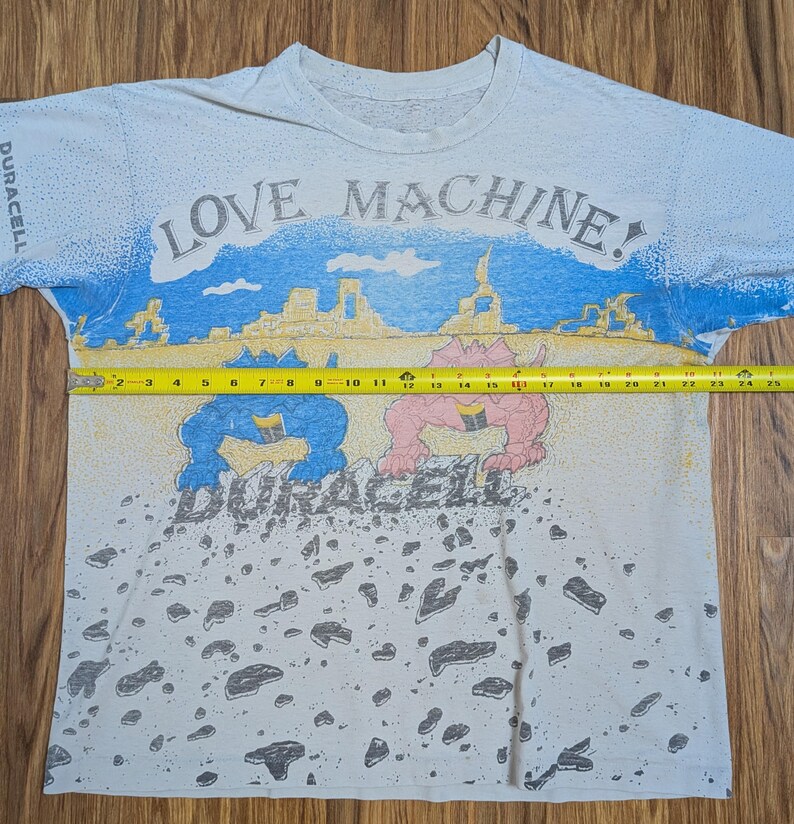 May include: White t-shirt with a colorful graphic design. The shirt features the text "LOVE MACHINE!" above a desert scene with cartoon figures and the Duracell logo. The shirt measures approximately 24 inches across.