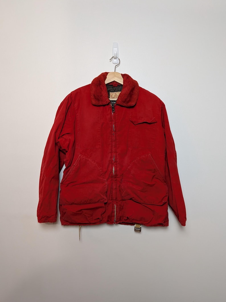 May include: A red corduroy jacket with a faux fur collar, a zip fastening, and several pockets. The jacket is hanging on a wooden hanger against a white wall. The jacket has a vintage appearance.