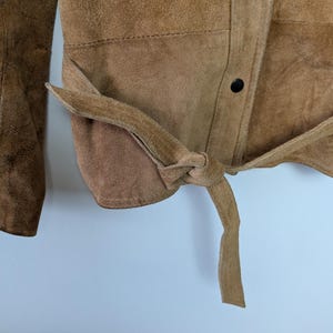 May include: A tan suede jacket featuring a tie-waist belt and a front placket with dark brown snap buttons. The garment has a textured surface and long sleeves, with the belt tied in a knot at the side to cinch the waist area of the jacket.