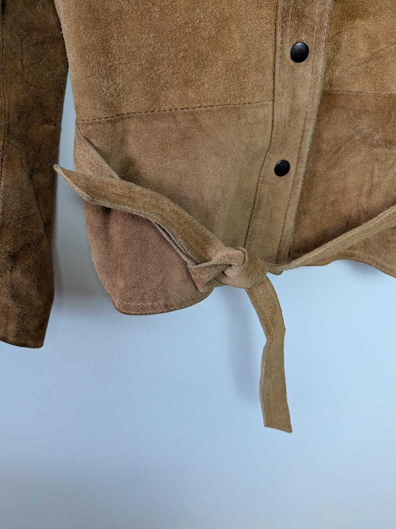 May include: A tan suede jacket featuring a tie-waist belt and a front placket with dark brown snap buttons. The garment has a textured surface and long sleeves, with the belt tied in a knot at the side to cinch the waist area of the jacket.