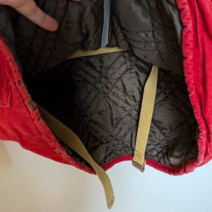 May include: A red quilted jacket interior with a dark brown lining. The jacket has beige straps and a zipper. The interior lining has a diamond pattern. The jacket is open, revealing the inside.