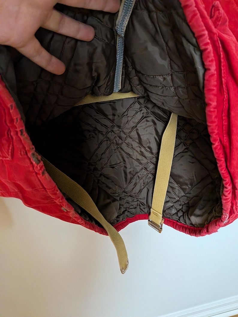 May include: A red quilted jacket interior with a dark brown lining. The jacket has beige straps and a zipper. The interior lining has a diamond pattern. The jacket is open, revealing the inside.