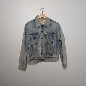 May include: Light blue denim jacket with a classic collar, button closure, and two chest pockets. The jacket shows signs of wear with distressed details and a faded appearance. It hangs from a wooden hanger against a plain white wall.