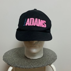 Könnte beinhalten: Schwarze Baseballkappe mit rosa gesticktem Text, der "bryan ADAMS" lautet.