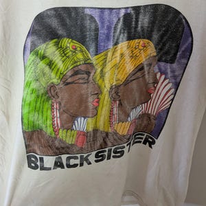 May include: Off-white t-shirt featuring a graphic of two women in profile, wearing headscarves and earrings. The women are depicted with brown skin, red lips, and holding fans. The text "BLACK SISTER" is printed below the image.
