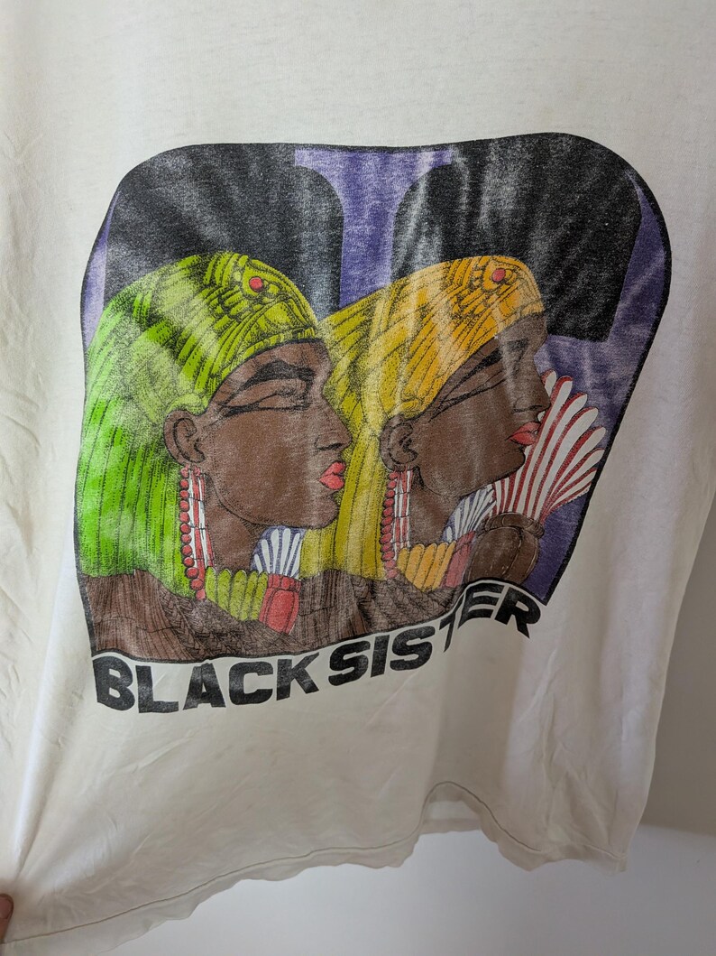 May include: Off-white t-shirt featuring a graphic of two women in profile, wearing headscarves and earrings. The women are depicted with brown skin, red lips, and holding fans. The text "BLACK SISTER" is printed below the image.