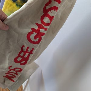 May include: A beige long-sleeve sweatshirt featuring the red text KIDS SEE GHOSTS printed vertically down the sleeve. The garment also displays a portion of a yellow and dark blue tie-dye pattern on the upper section of the fabric.