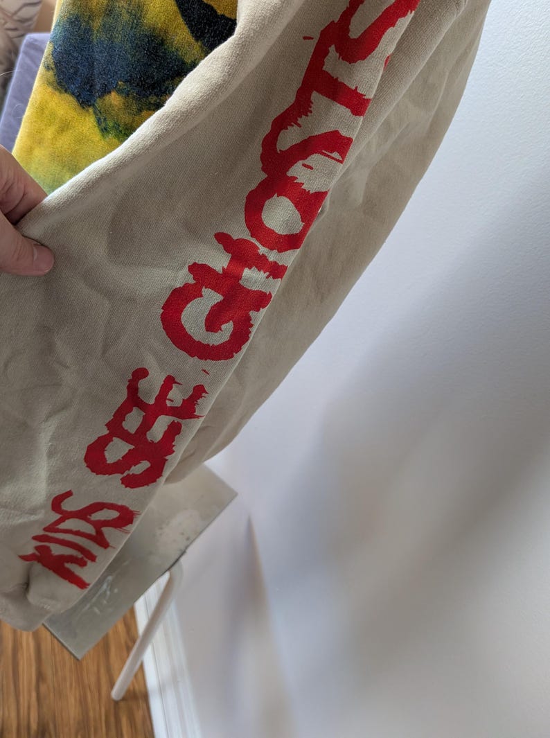 May include: A beige long-sleeve sweatshirt featuring the red text KIDS SEE GHOSTS printed vertically down the sleeve. The garment also displays a portion of a yellow and dark blue tie-dye pattern on the upper section of the fabric.