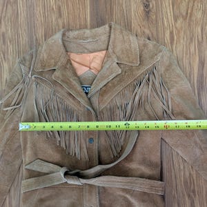 May include: A tan suede jacket featuring long fringe detailing across the chest and shoulders, a collared neckline, and a tie-waist belt. A yellow measuring tape is placed across the front, showing a width of approximately 19 inches.