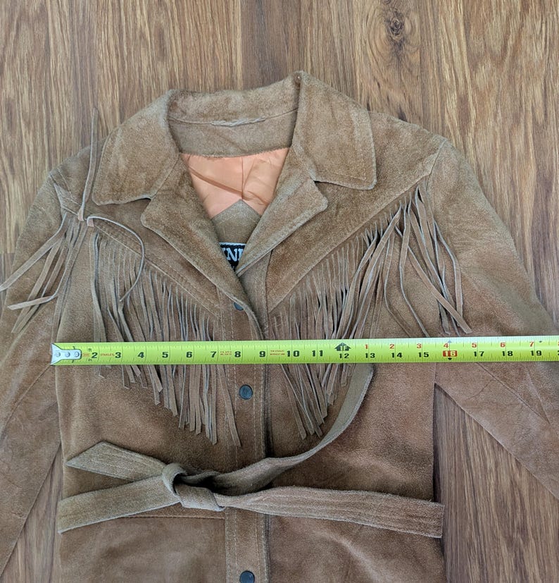 May include: A tan suede jacket featuring long fringe detailing across the chest and shoulders, a collared neckline, and a tie-waist belt. A yellow measuring tape is placed across the front, showing a width of approximately 19 inches.