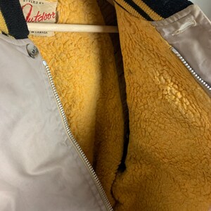 May include: A beige bomber jacket with a black and gold striped collar and a zipper closure. The jacket is lined with a soft, yellow fleece material. The label on the jacket reads "Styled by Outdoor, Made in Canada."