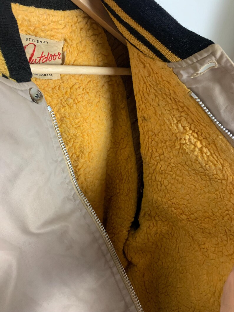May include: A beige bomber jacket with a black and gold striped collar and a zipper closure. The jacket is lined with a soft, yellow fleece material. The label on the jacket reads "Styled by Outdoor, Made in Canada."