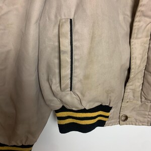 May include: A beige bomber jacket with black and yellow striped trim. The jacket has a zipper closure and a pocket on the left side.