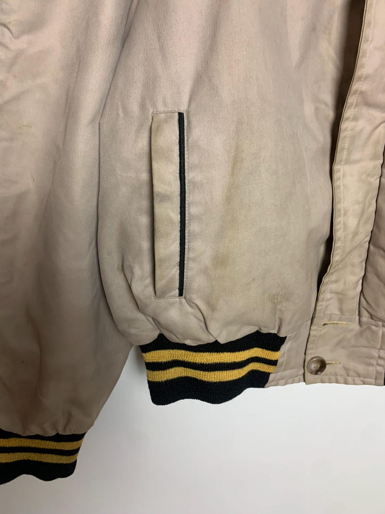 May include: A beige bomber jacket with black and yellow striped trim. The jacket has a zipper closure and a pocket on the left side.