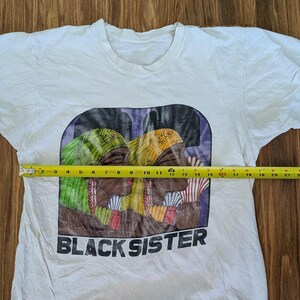 May include: White t-shirt with a graphic print featuring two women with colorful head wraps and the text "BLACK SISTER" in black. The graphic measures approximately 10 inches wide.