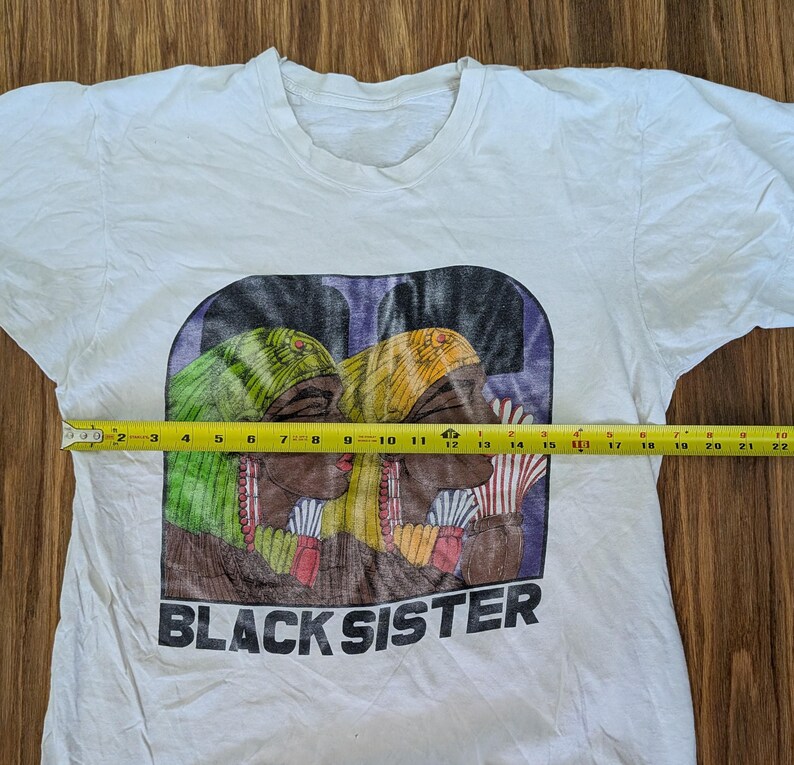 May include: White t-shirt with a graphic print featuring two women with colorful head wraps and the text "BLACK SISTER" in black. The graphic measures approximately 10 inches wide.