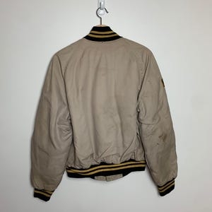 May include: A beige bomber jacket with black and gold striped ribbing around the collar, cuffs, and hem. The jacket has a patch on the right sleeve.