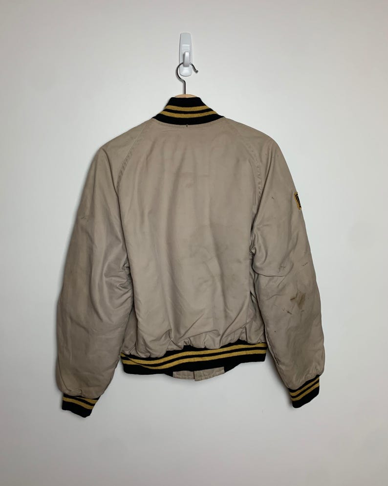 May include: A beige bomber jacket with black and gold striped ribbing around the collar, cuffs, and hem. The jacket has a patch on the right sleeve.