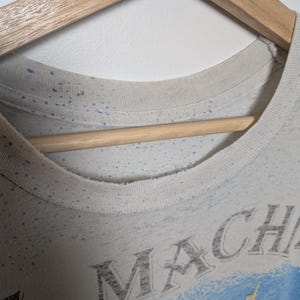 May include: Close-up of a light-colored t-shirt with a crew neck, hanging on a wooden hanger. The shirt has a faded design with the word "MACH" visible. Blue speckles are scattered across the fabric.