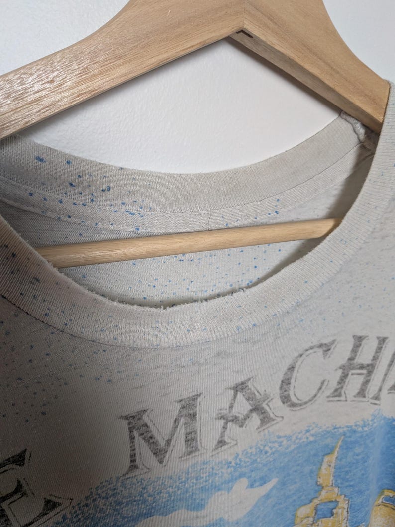 May include: Close-up of a light-colored t-shirt with a crew neck, hanging on a wooden hanger. The shirt has a faded design with the word "MACH" visible. Blue speckles are scattered across the fabric.