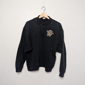 Peut inclure: Sweat-shirt noir à manches longues avec un col et une patte de boutonnage à trois boutons. Le sweat-shirt présente un motif brodé sur la poitrine avec le mot "BACADRAFT" au-dessus d'un emblème circulaire avec des accents rouges, jaunes et blancs.