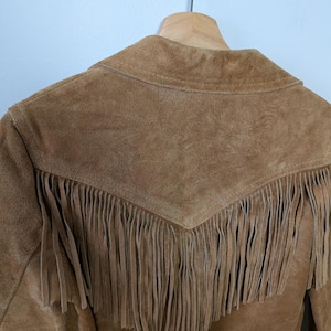 May include: A tan suede jacket featuring a pointed back yoke with long, hanging fringe detailing. The garment is constructed from a textured leather material and includes a collar, hanging from a wooden hanger against a plain wall.