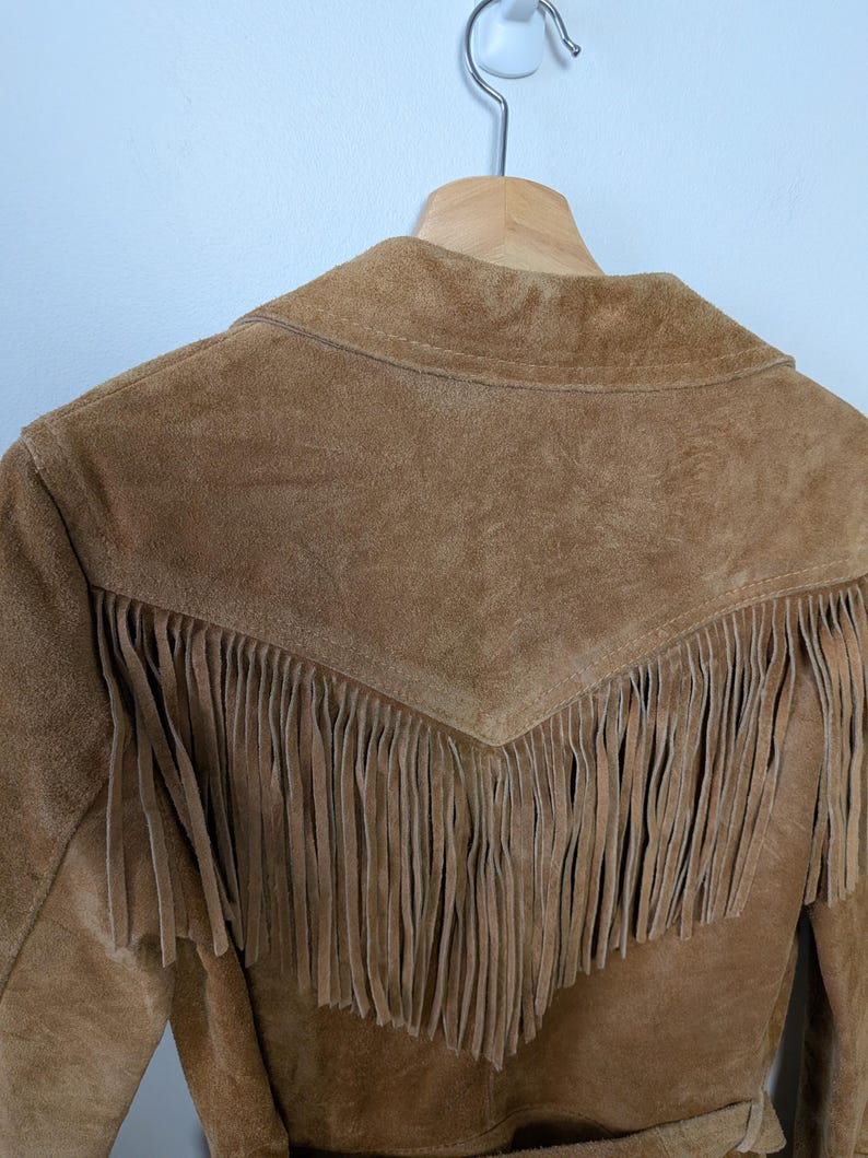 May include: A tan suede jacket featuring a pointed back yoke with long, hanging fringe detailing. The garment is constructed from a textured leather material and includes a collar, hanging from a wooden hanger against a plain wall.