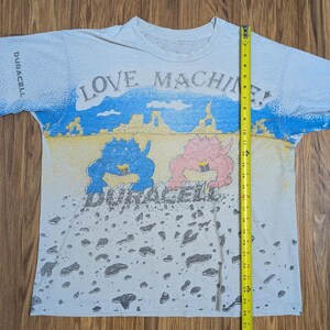 May include: White t-shirt with a colorful graphic design. The shirt features the text "LOVE MACHINE!" and "DURACELL". The design includes blue and pink dinosaur-like creatures and a landscape with blue sky and yellow sand. The shirt is approximately 27 inches long.