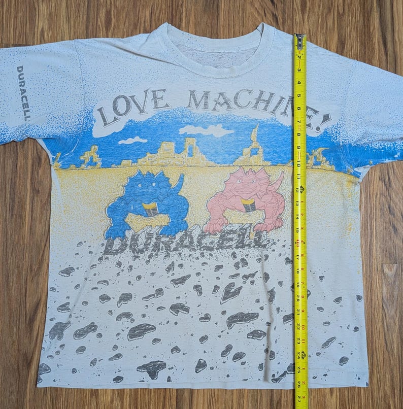 May include: White t-shirt with a colorful graphic design. The shirt features the text "LOVE MACHINE!" and "DURACELL". The design includes blue and pink dinosaur-like creatures and a landscape with blue sky and yellow sand. The shirt is approximately 27 inches long.