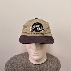 Può includere: Un cappellino da baseball color beige con visiera blu scuro. La parte anteriore presenta un cerchio blu scuro ricamato con stelle bianche e la scritta PLANET HOLLYWOOD in bianco, con le parole San Francisco ricamate in nero sotto il cerchio.