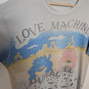May include: Vintage gray t-shirt with the words "LOVE MACHINE" printed across the top. The shirt features a colorful graphic of two cartoon dinosaurs in a desert landscape. The shirt is hanging on a wooden hanger.
