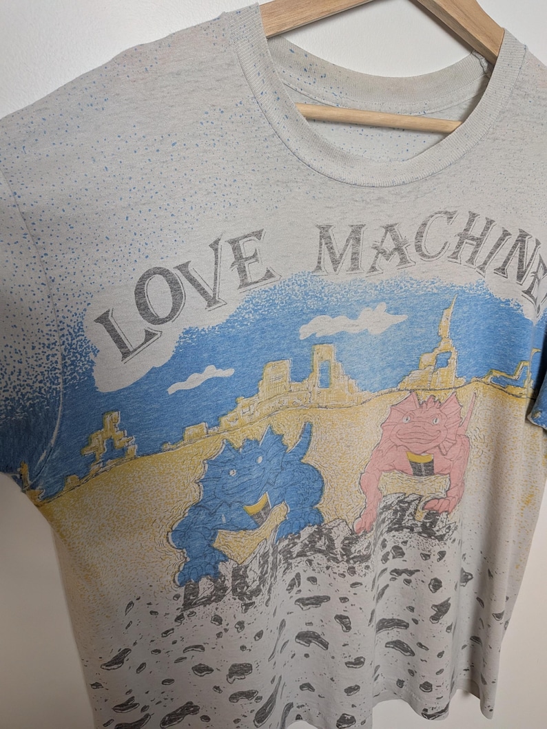 May include: Vintage gray t-shirt with the words "LOVE MACHINE" printed across the top. The shirt features a colorful graphic of two cartoon dinosaurs in a desert landscape. The shirt is hanging on a wooden hanger.