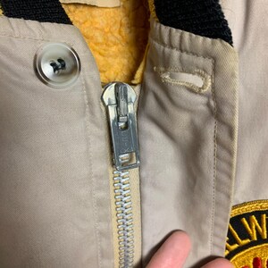 May include: Close-up of a beige jacket with a zipper and a yellow and black patch that says "All-Weather Club". The jacket has a black and yellow collar and a yellow fleece lining.