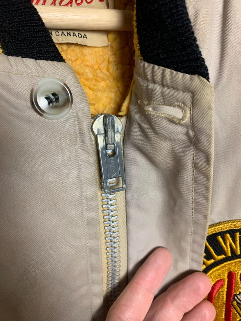 May include: Close-up of a beige jacket with a zipper and a yellow and black patch that says "All-Weather Club". The jacket has a black and yellow collar and a yellow fleece lining.