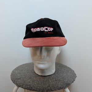 Può includere: Cappellino da baseball nero Robocop The Series con visiera rossa. Il testo "ROBOCOP THE SERIES" è ricamato in rosso e bianco sul davanti. Il cappellino è esposto su una testa di manichino bianca.