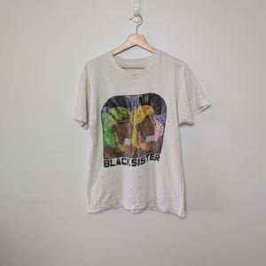 May include: Off-white t-shirt with a graphic print featuring two women's profiles with colorful head wraps. The text "BLACK SISTER" is printed below the image. The shirt is hanging on a wooden hanger against a white wall.