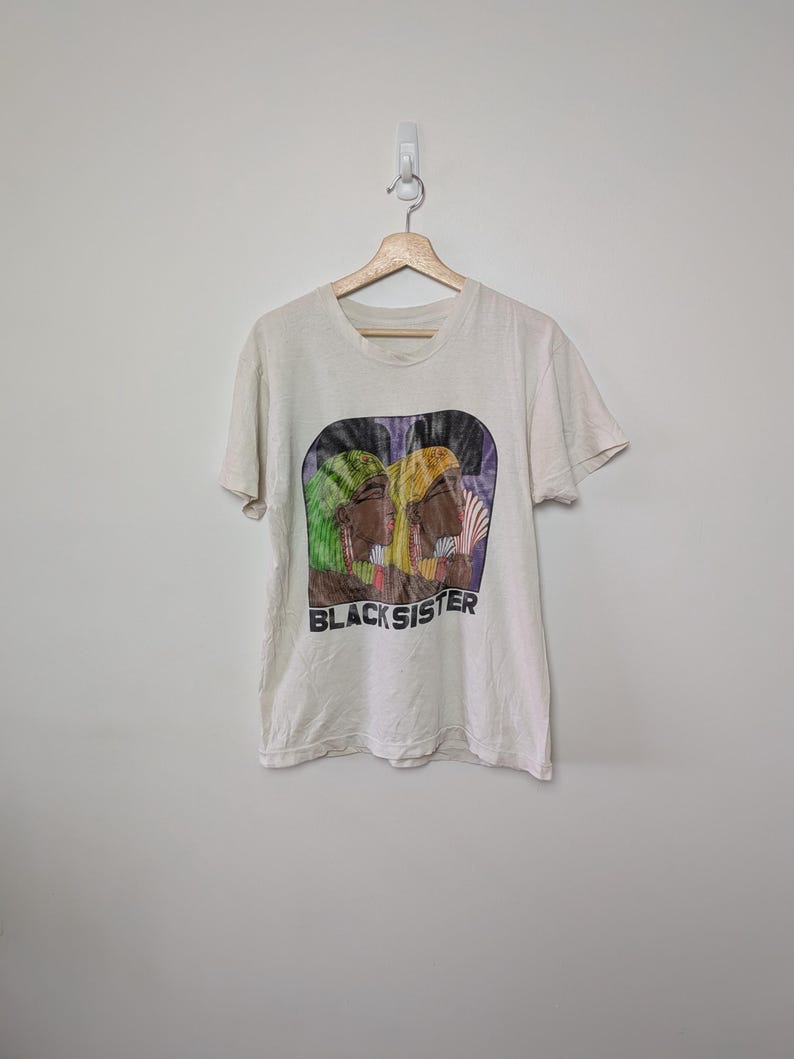 May include: Off-white t-shirt with a graphic print featuring two women's profiles with colorful head wraps. The text "BLACK SISTER" is printed below the image. The shirt is hanging on a wooden hanger against a white wall.