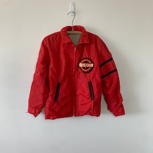 May include: Red nylon jacket with black stripes on the sleeves and a black and orange patch that says "Technical" and "UNB" on the front. The jacket has a zipper closure and two pockets.