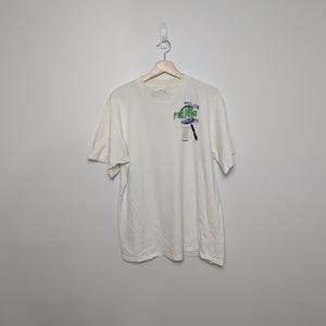 Puede incluir: Camiseta de color crema con un gráfico en la parte delantera. El gráfico incluye las palabras "PAJARO" y "DISCOVERY" en verde y azul, junto con una ilustración de una lupa. La camiseta está colgada en una percha de madera.