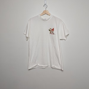 May include: White t-shirt with a small graphic on the front. The graphic features a colourful illustration of a person with a red hat and other figures. The t-shirt is hanging on a wooden hanger.