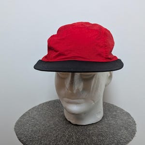 May include: A red and black bucket hat. The hat features a red crown and a black brim. The hat is displayed on a white mannequin head, against a neutral background. This hat is a fashionable accessory.