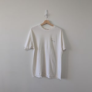 以下が含まれることがあります： 木製のハンガーに掛けられたオフホワイトのTシャツ。シャツの左胸上部に小さなグラフィックとテキストがあります。シャツは柔らかく、軽量な素材で作られています。