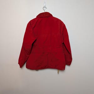 May include: A red jacket with a corduroy collar hangs on a wall hook. The jacket has a button closure, pockets, and a waist belt. The jacket is a solid red colour.
