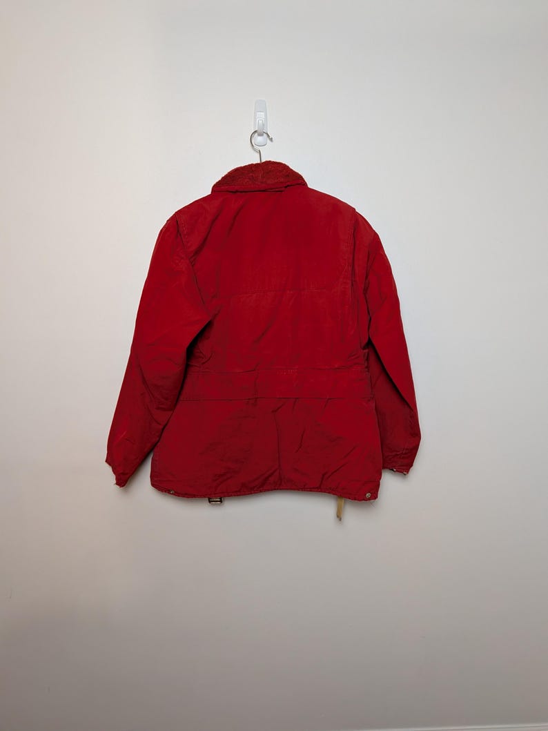 May include: A red jacket with a corduroy collar hangs on a wall hook. The jacket has a button closure, pockets, and a waist belt. The jacket is a solid red colour.