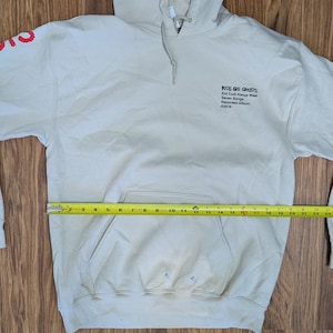 May include: A beige hooded sweatshirt with a front kangaroo pocket and a yellow measuring tape across the midsection showing a width of 24 inches. The chest features black text reading Kids See Ghosts, and the sleeve has red graphic lettering.