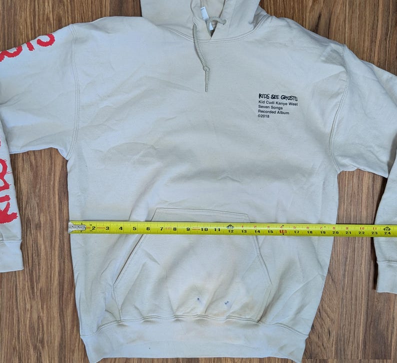 May include: A beige hooded sweatshirt with a front kangaroo pocket and a yellow measuring tape across the midsection showing a width of 24 inches. The chest features black text reading Kids See Ghosts, and the sleeve has red graphic lettering.