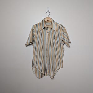 May include: A short-sleeved shirt with vertical stripes in brown, yellow, and pale blue. The shirt has a single chest pocket and is hanging on a wooden hanger against a plain white wall.