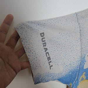 May include: White t-shirt sleeve with a blue and yellow design. The sleeve has a speckled pattern and the word "DURACELL" printed in gray.