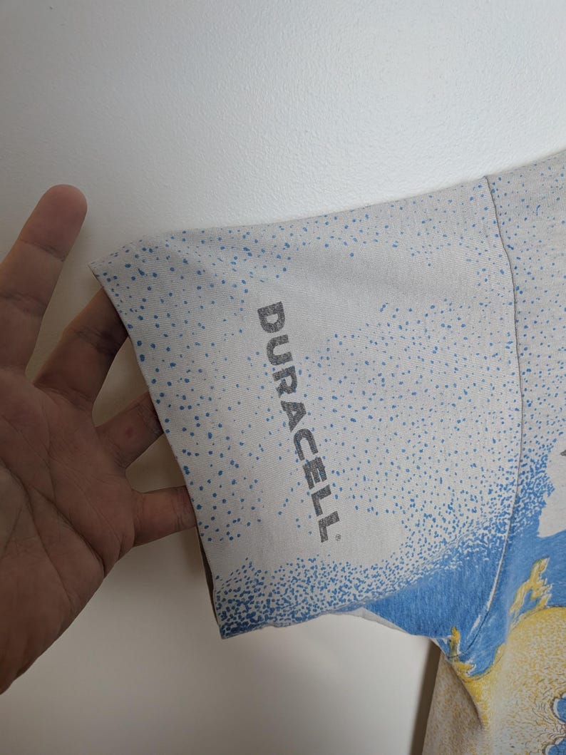 May include: White t-shirt sleeve with a blue and yellow design. The sleeve has a speckled pattern and the word "DURACELL" printed in gray.