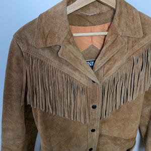 May include: A tan suede jacket featuring long fringe detailing across the chest and shoulders. The jacket has a classic collar, a button-front closure with black buttons, and an orange interior lining. A partial brand label is visible at the neck.